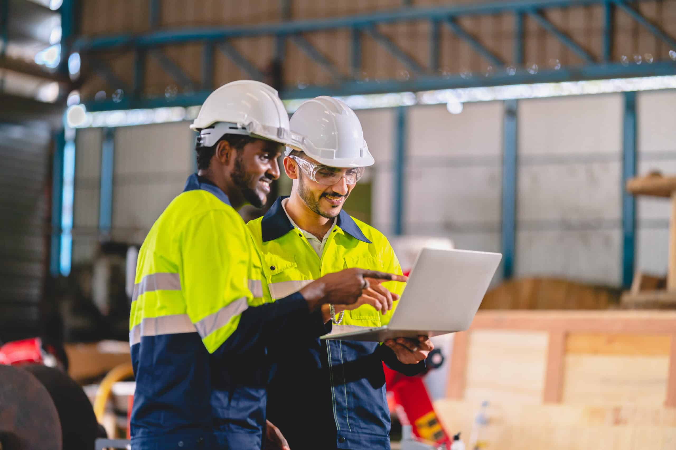 Workers in safety gear review turnaround mechanical services on a laptop inside an industrial warehouse.