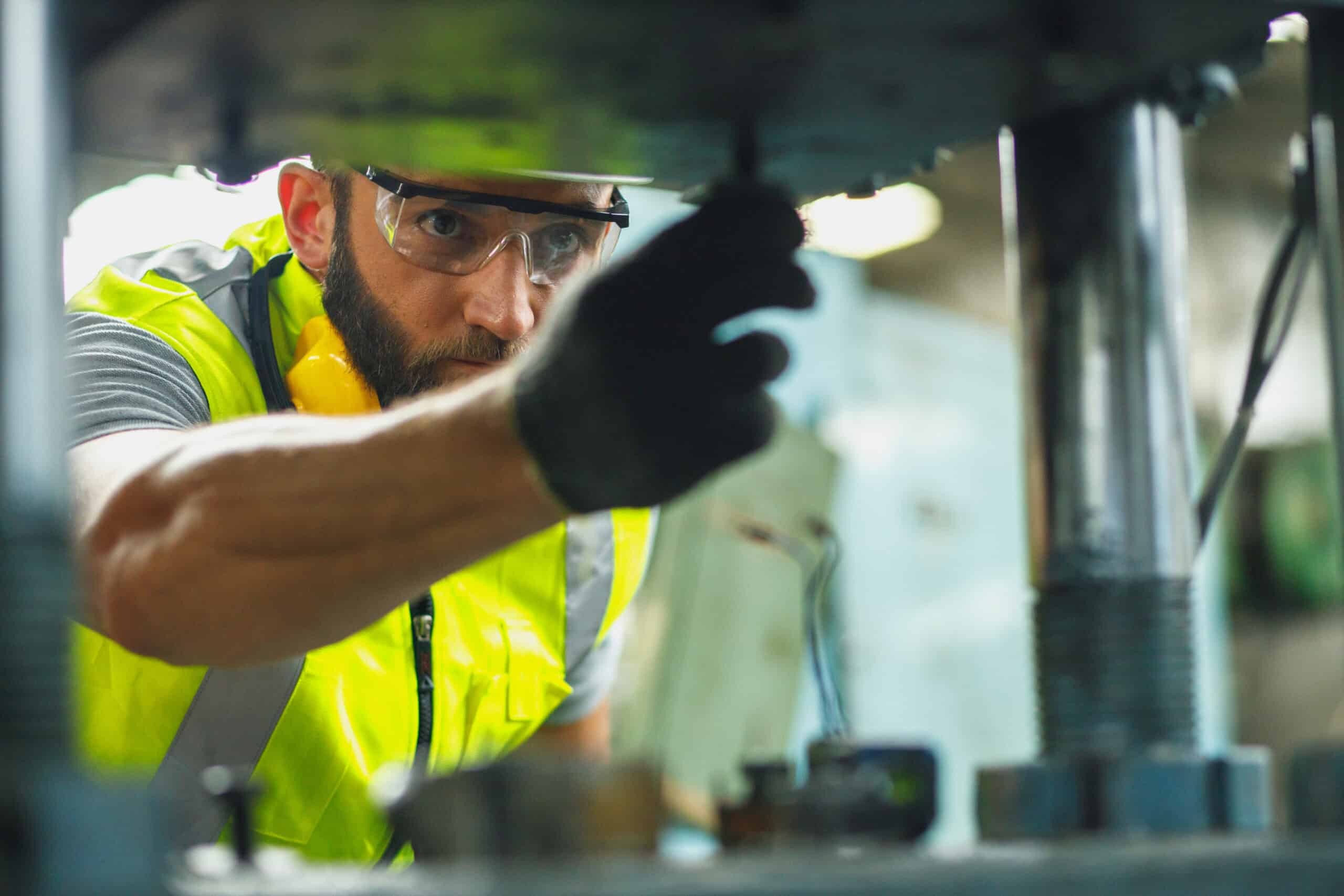 A worker in a yellow safety vest and goggles adjusts machinery, performing industrial maintenance.