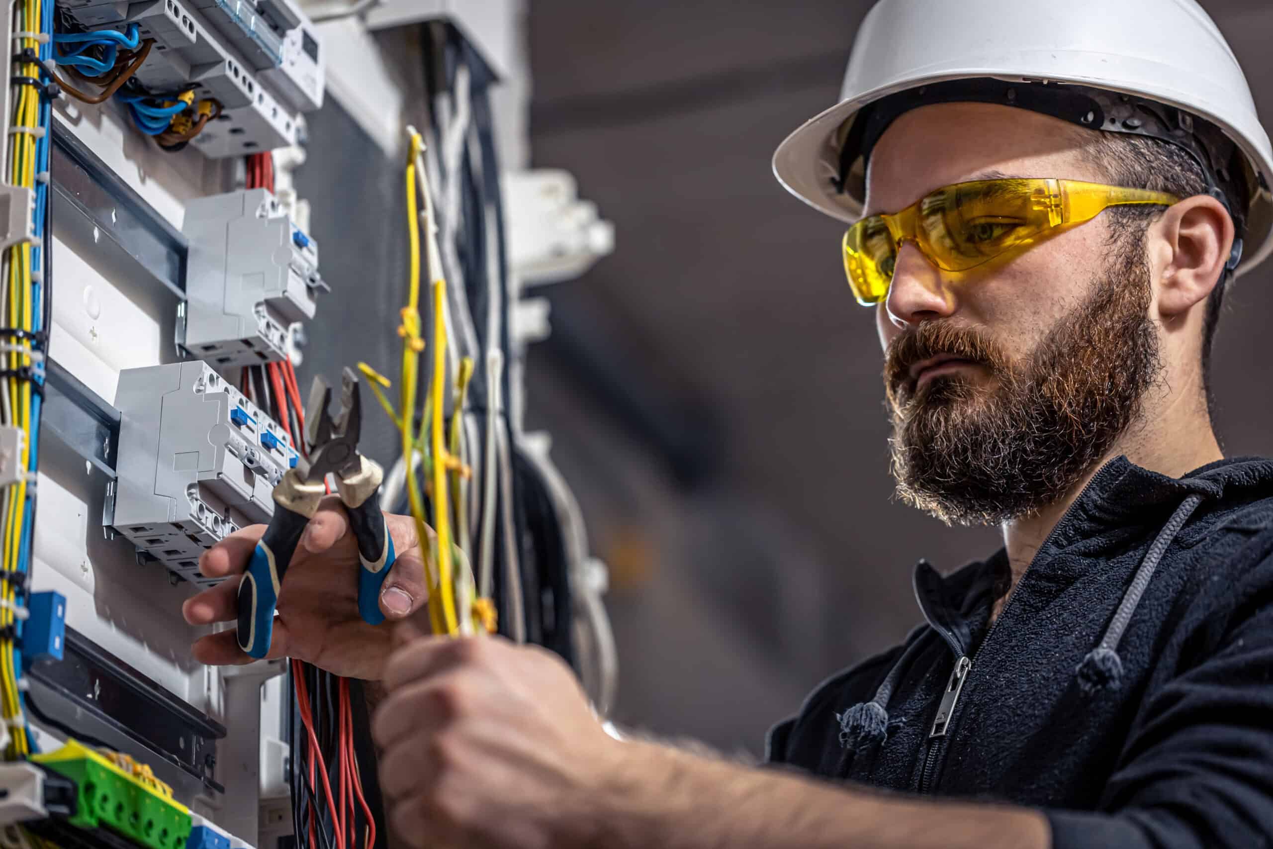 Bearded electrician in white hard hat and yellow safety glasses works on electrical wiring, showing reliability.