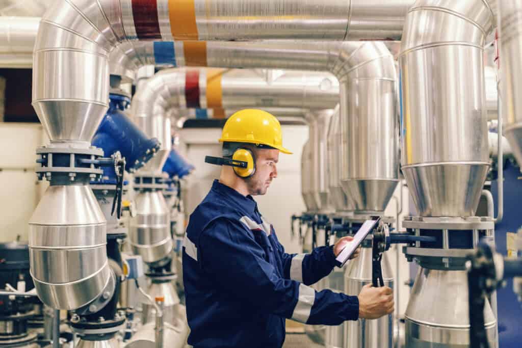 Worker in yellow hard hat and ear protection checks clipboard while inspecting equipment during industrial maintenance.