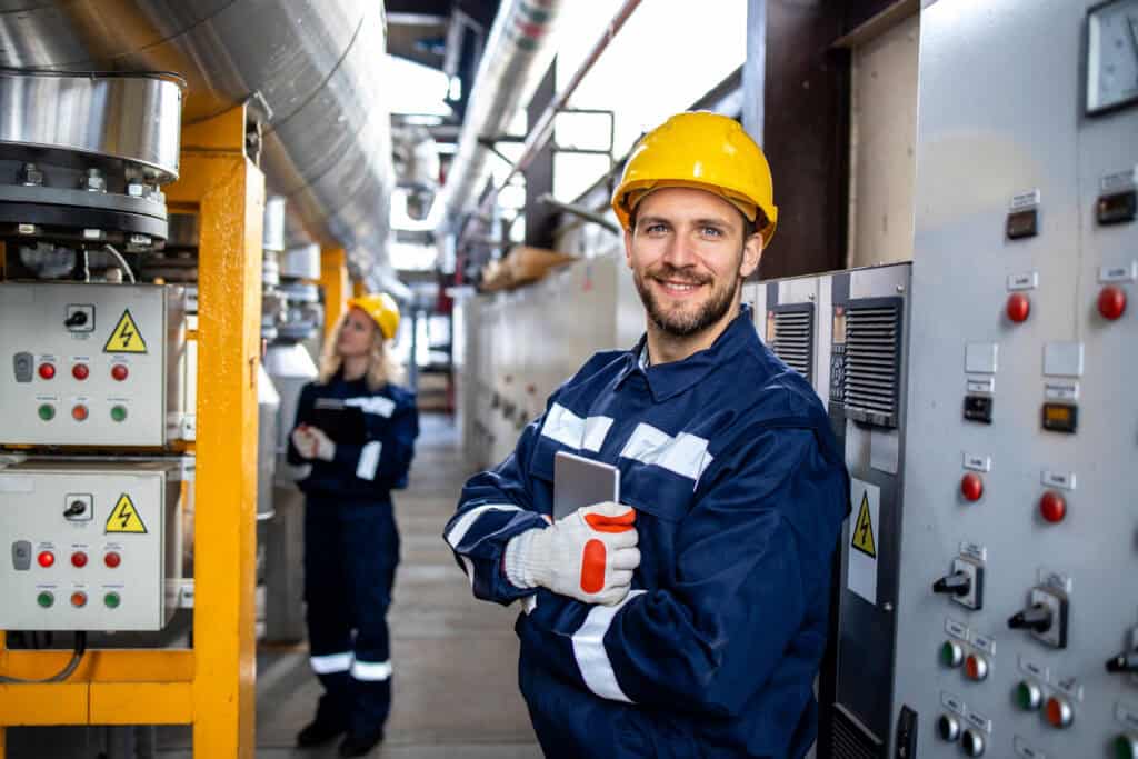 Smiling worker in hard hat stands in industrial facility, highlighting maintenance expertise; coworker visible behind.